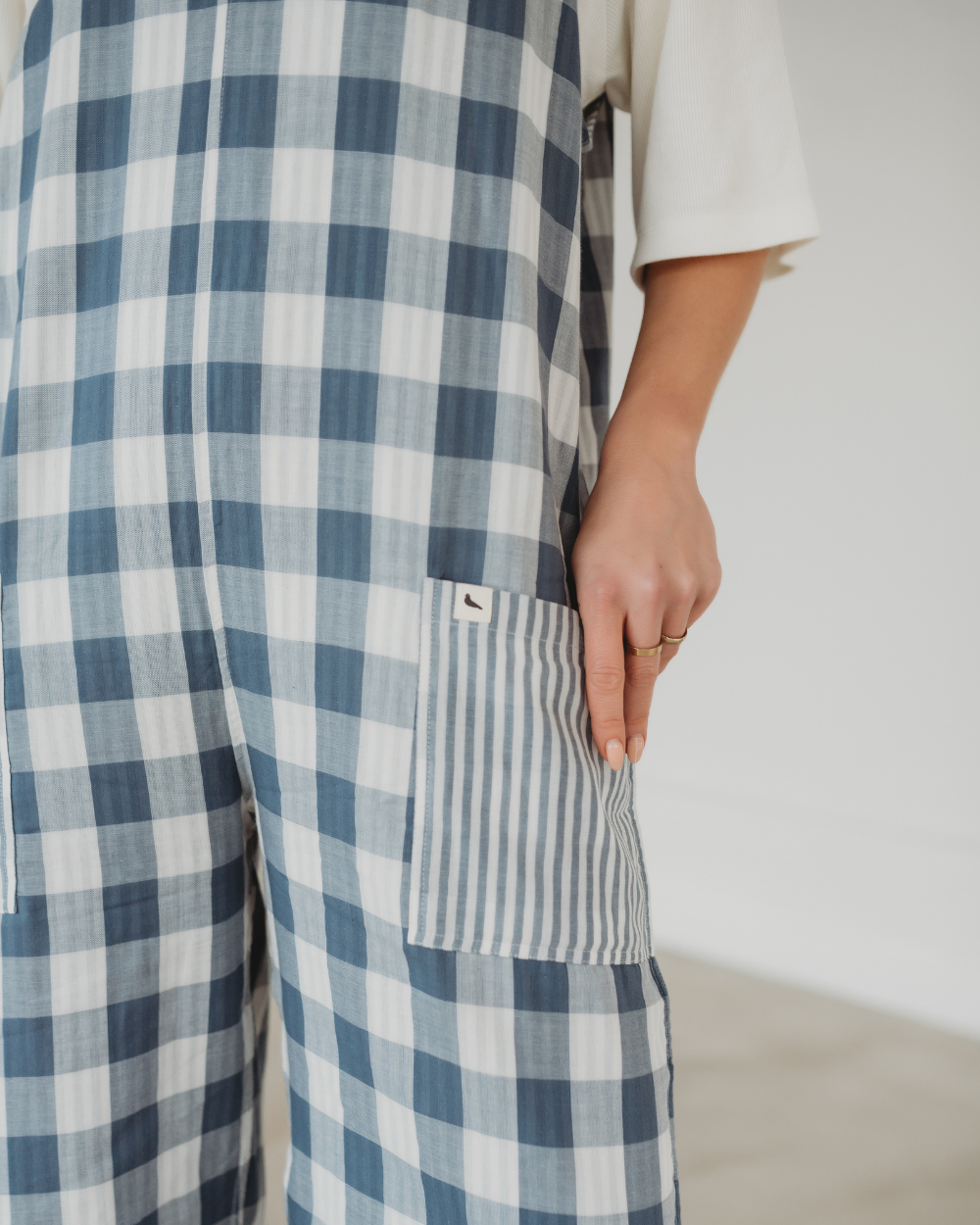 Someone wears Grace Reversible Blue Check Dungarees—crafted from organic cotton—with a large striped pocket, hand resting on it. A blurred background highlights the sustainable design and texture of the blue and white checkered jumpsuit.