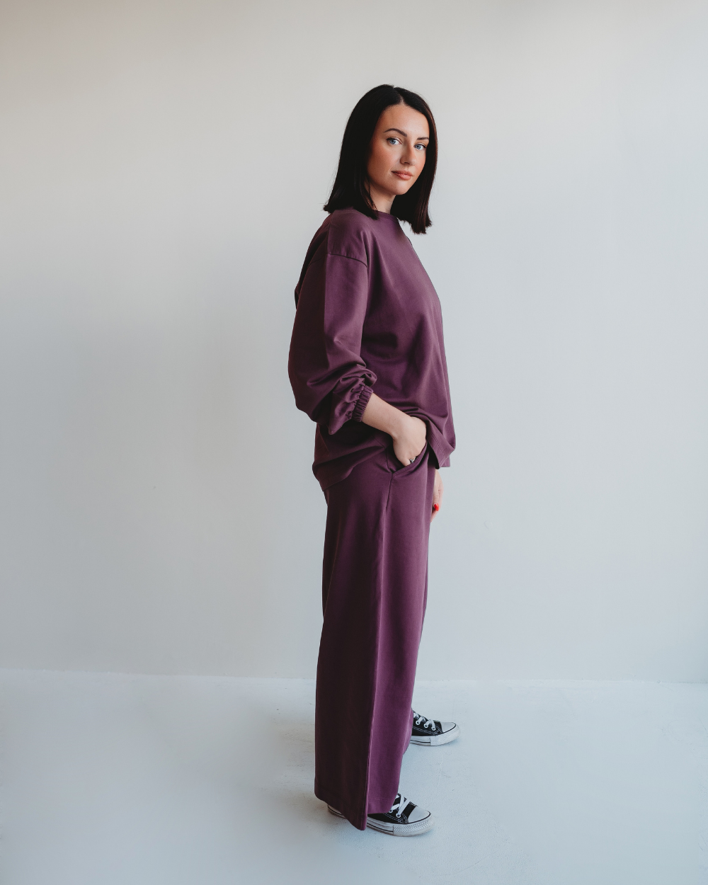 A woman with shoulder-length dark hair poses against a light background, wearing Noor Wide Leg Plum Trousers and a matching maroon sweatshirt. With one hand in her pocket and stylish black and white sneakers, she gazes at the camera.