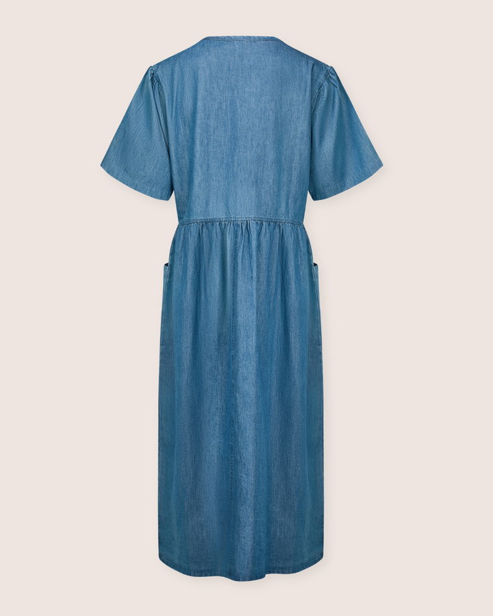 The Magda Washed Denim Blue Dress, displayed from the back, features short sleeves and a V-neck. Made from organic cotton, it has a gathered waist and straight hemline for a casual look. The smooth denim texture is striking against the plain off-white background.