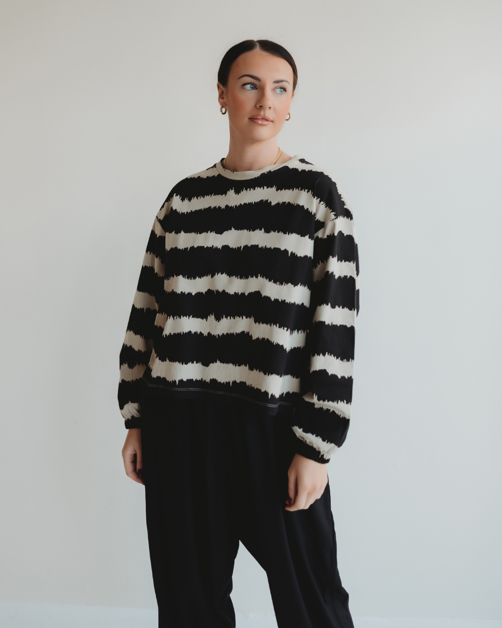 A person stands against a plain background wearing the Ivy Drift Print Top—a black and white striped sweater with a wavy pattern and balloon sleeves, crafted from organic cotton. They pair it with black pants, dark hair pulled back, gazing slightly to the side.