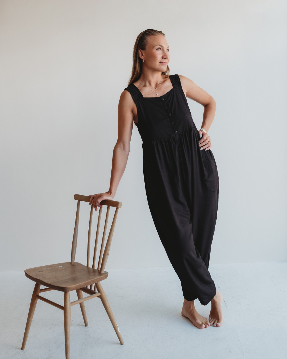 A woman in the Alice Black Viscose Jumpsuit, crafted from eco viscose, leans against a wooden chair barefoot on a light-colored floor, gazing thoughtfully to the side against a plain white background.