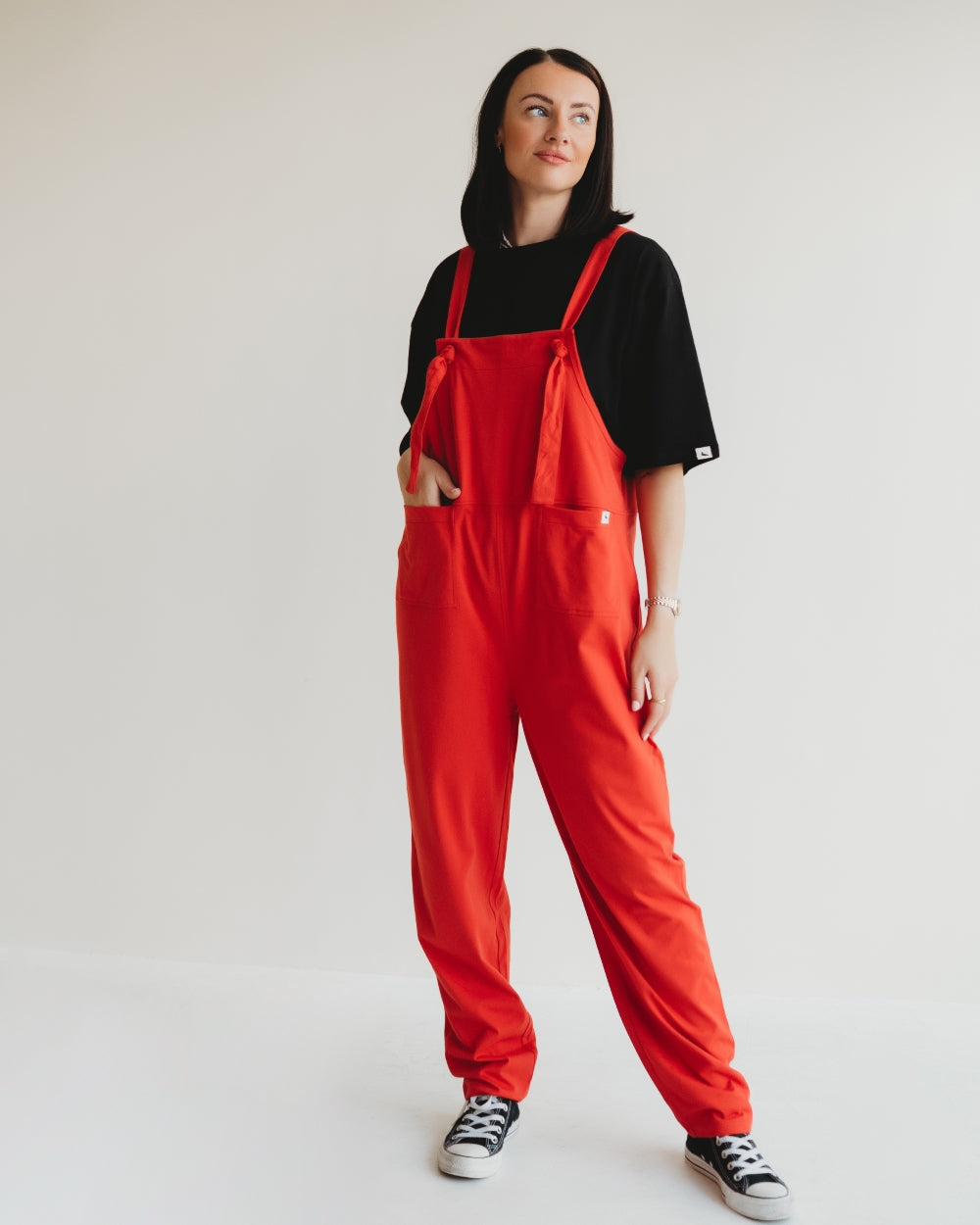 Against a white background, a conscious consumer wears Lila Plain Red Dungarees over an organic cotton black t-shirt. Complemented by black and white sneakers, their hands rest casually in the pockets as they gaze slightly to the side.