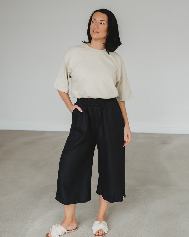 In a minimalist room, a person with dark hair wears Judy Black Twill Trousers made from organic cotton, featuring an elastic waistband, paired with a light-colored t-shirt and white slide sandals. They gaze slightly to the side with one hand in their pocket.