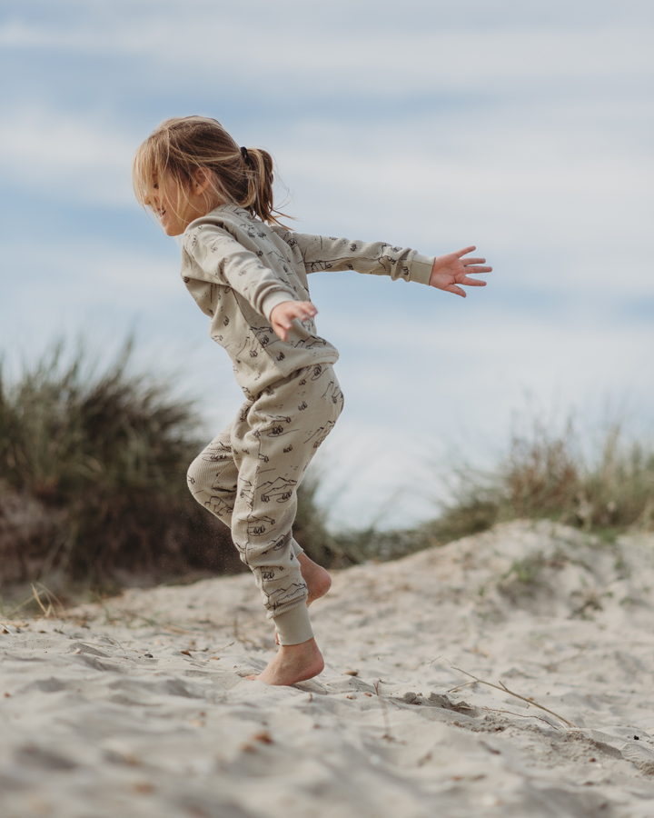 A child with long hair joyfully leaps on a sandy beach dotted with grass tufts, wearing Wilderness Joggers and a printed beige long-sleeve top. The partly cloudy sky adds to the serene and playful scene.