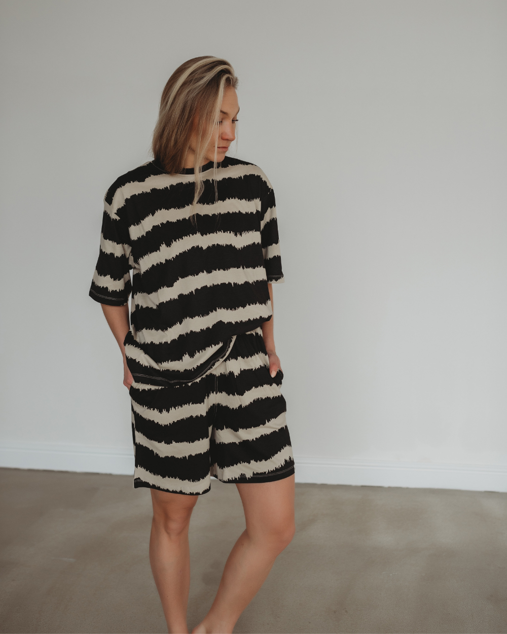 A person in a matching black and white patterned shirt and Audrey Drift Print Shorts, crafted from organic cotton, stands barefoot indoors against a plain white wall. Their hands rest in pockets as they gaze to the side, showcasing a sustainable wardrobe choice.