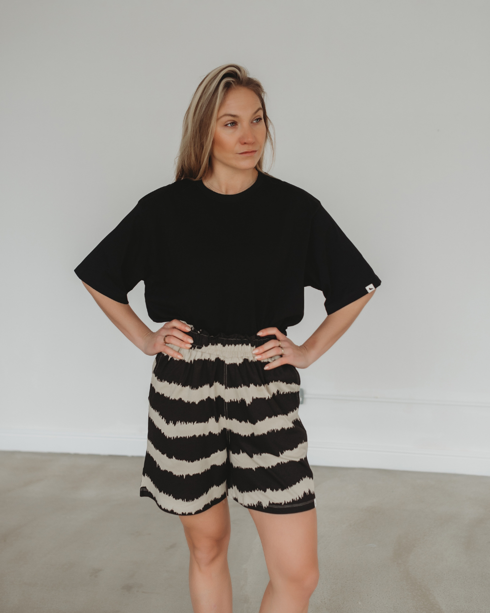 Against a plain background, a person stands confidently with hands on hips, wearing an organic cotton black T-shirt and Audrey Drift Print Shorts. The expression is thoughtful in this minimalistic setting.