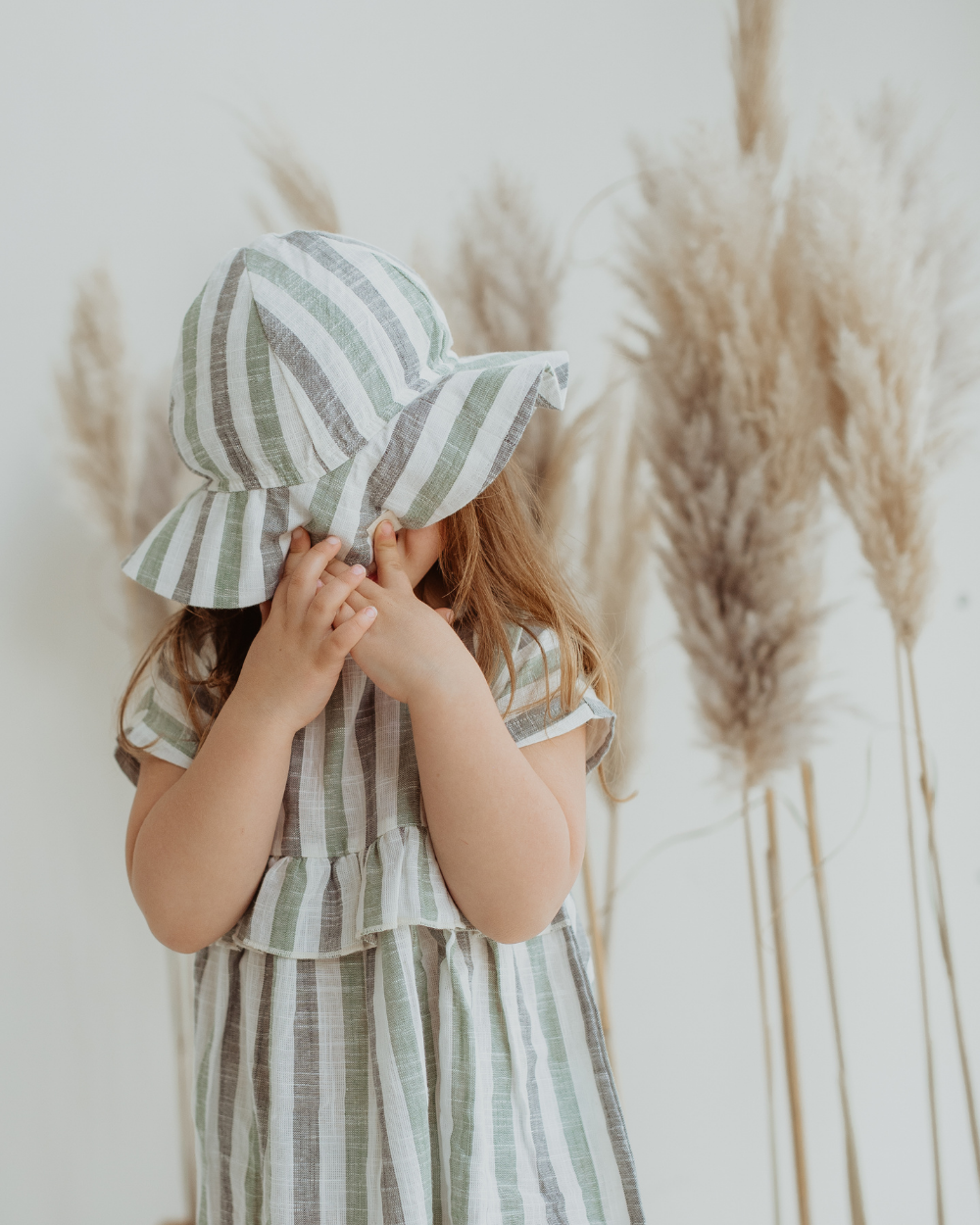 A child with their hands covering their face wearing a beige and green striped sun hat and a matching dress.