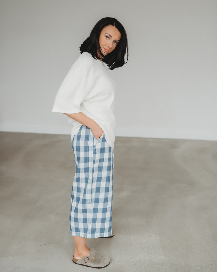 A person with shoulder-length black hair stands in a minimalistic room, wearing a white top, Judy Reversible Blue Check Trousers with an elasticated waistband, and brown slip-on shoes. The room features a plain gray floor and light-colored walls.