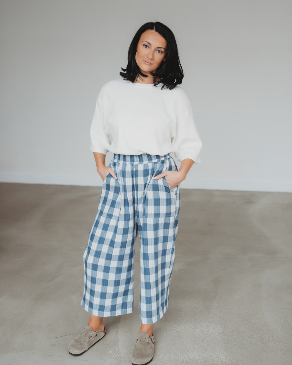 A black-haired woman poses indoors in a white sweater and Judy Reversible Blue Check Trousers, with her hands in her pockets. She stands on a light gray floor, set against a plain white wall.