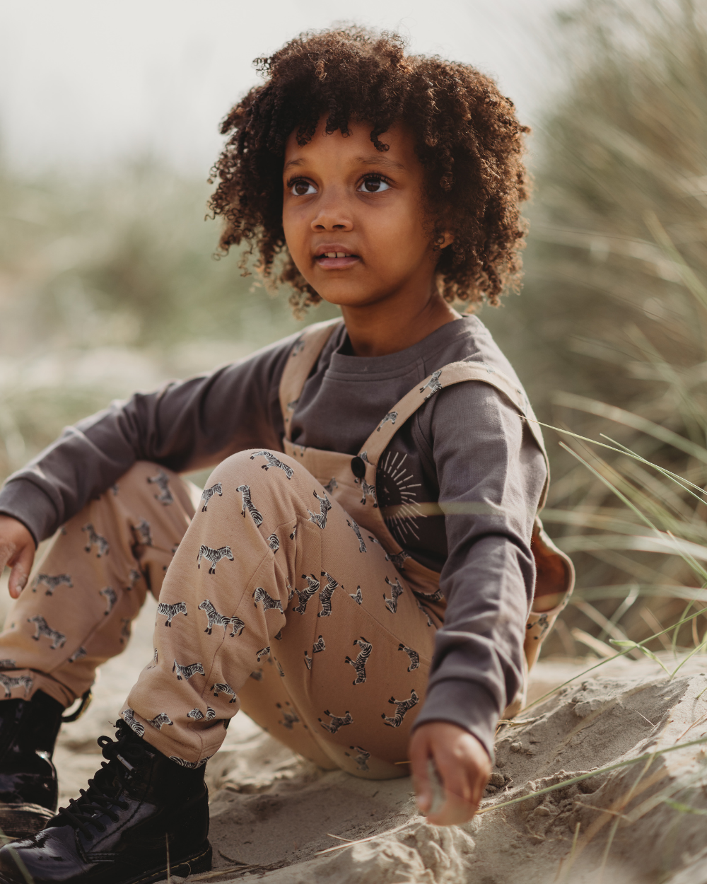 A young child with curly hair sits on sandy ground, wearing a gray eco-friendly long-sleeve shirt and animal-printed tan Zebra Dungarees. Black boots complete the look. They gaze thoughtfully to the side, surrounded by grassy plants.