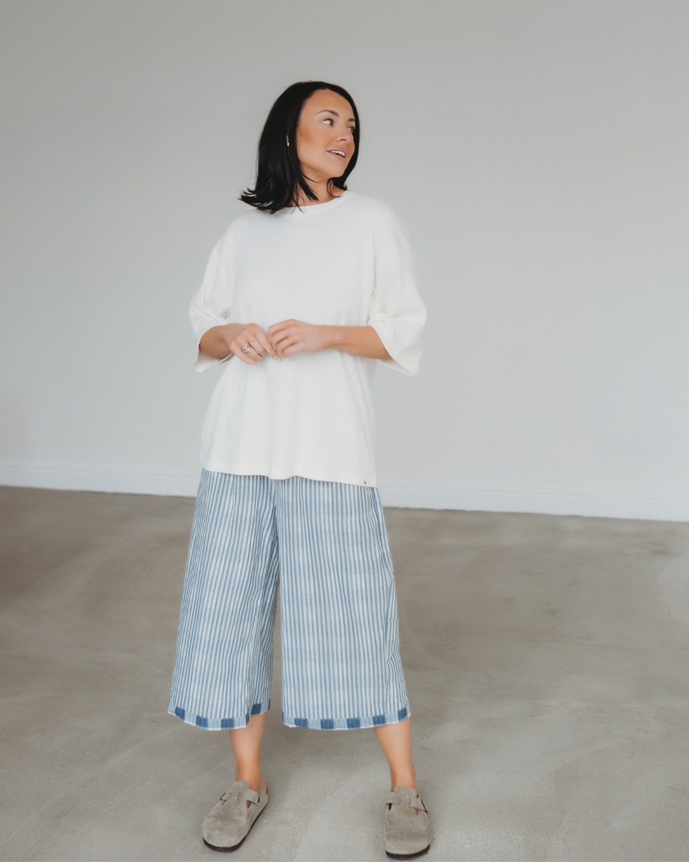 A person with shoulder-length dark hair wears a loose white top and Judy Reversible Blue Check Trousers made from organic cotton, featuring an elasticated waistband. Theyre paired with gray slip-on shoes, standing on a plain concrete floor against a neutral wall.