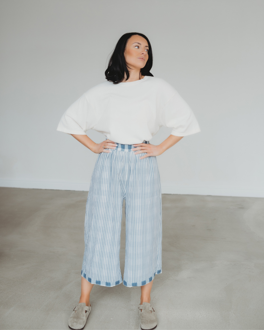 A woman stands confidently indoors, hands on hips, wearing a white blouse and the Judy Reversible Blue Check Trousers with an elasticated waistband, paired with beige shoes. The rooms decor features a plain white wall and light gray flooring.