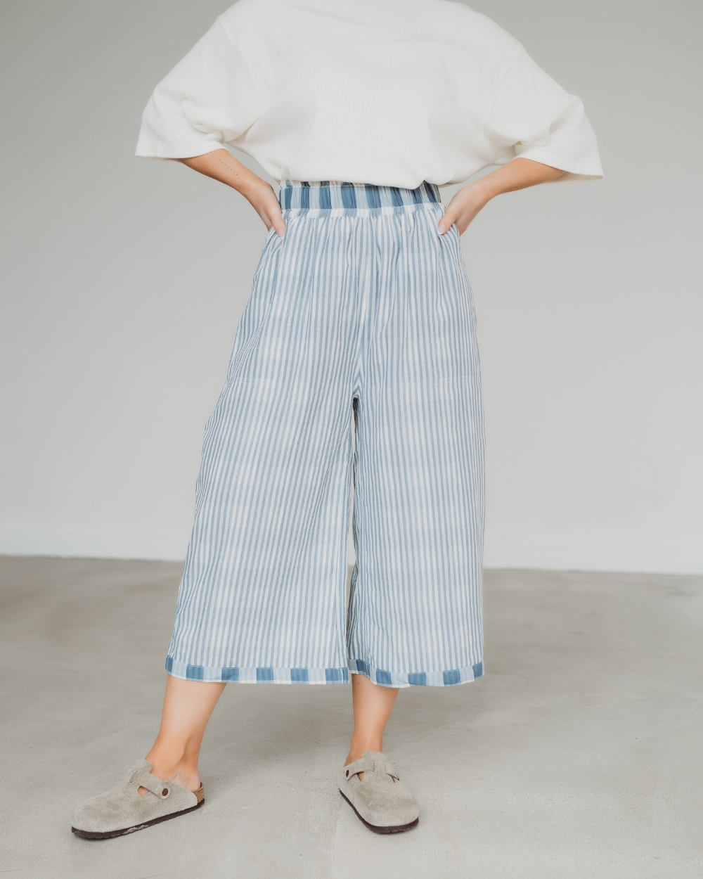 A person stands with hands on hips, wearing Judy Reversible Blue Check Trousers featuring light blue stripes and an elasticated waistband. Theyre paired with a white top and beige slip-on shoes on a light-colored floor against a plain wall.