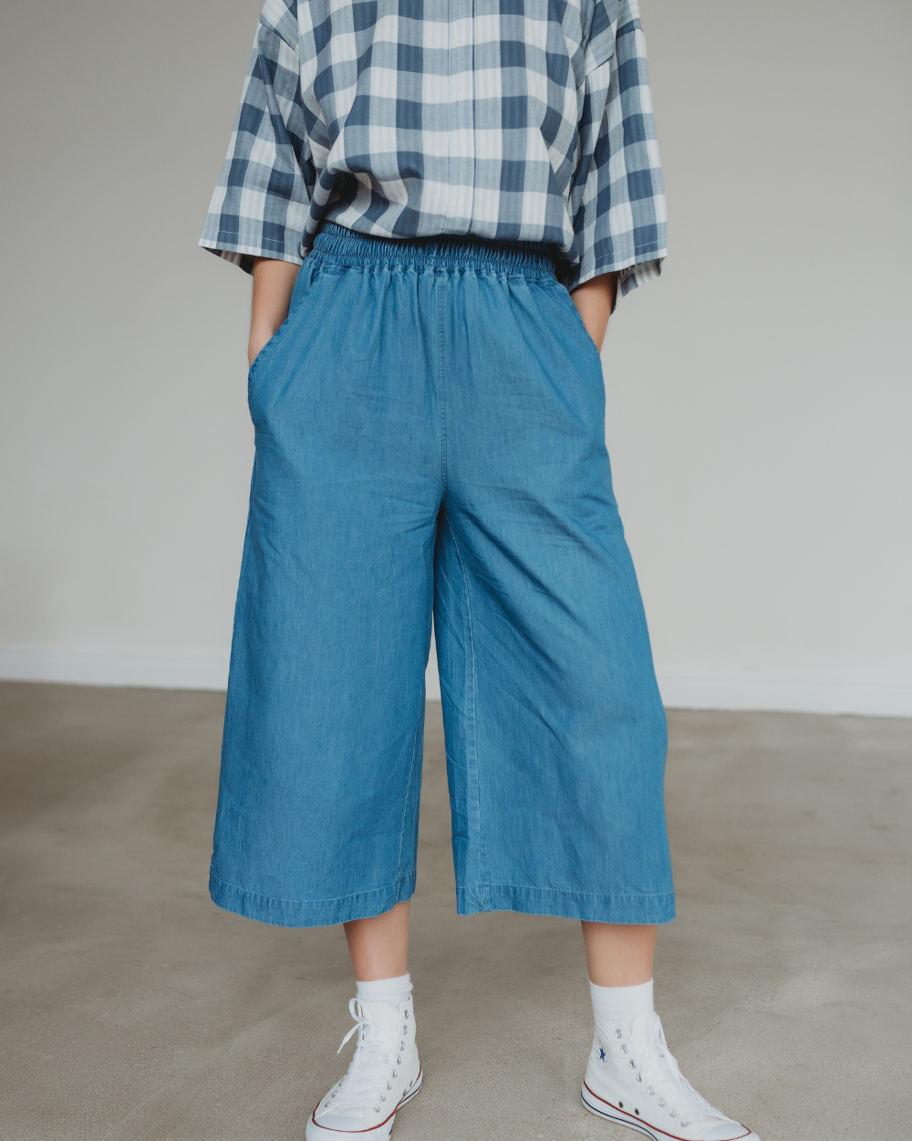 A person is wearing a blue and white plaid shirt tucked into Judy Washed Denim Blue Trousers made from organic cotton. Their hands rest in their pockets, standing on a gray floor in white high-top sneakers. The head is out of frame, emphasizing the comfort of an elasticated waistband.