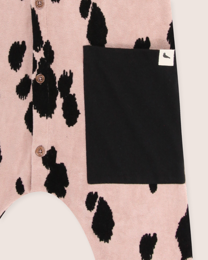 Close-up of Pony Velour Dungarees made from organic velour fabric, featuring a pink background with black cow-like spots. The garment includes brown buttons and a large black pocket with a small white tag, ideal for sustainable wardrobes.