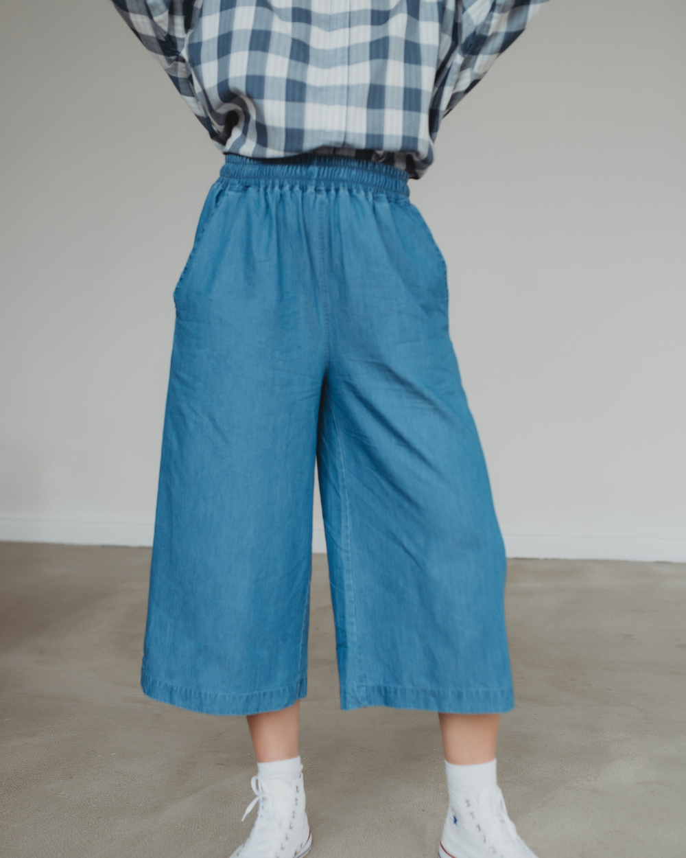 A person in a plaid shirt and Judy Washed Denim Blue Trousers with an elasticated waistband stands casually, hands in pockets. White sneakers complete the look against a plain, light-colored wall and floor backdrop.