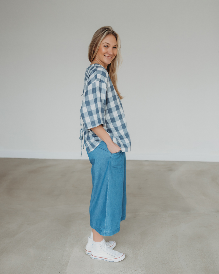 A person with long hair is smiling and standing sideways in a minimalistic indoor space, wearing a blue and white checkered top, Judy Washed Denim Blue Trousers with an elastic waistband, and white sneakers. The plain background highlights their effortlessly chic style.