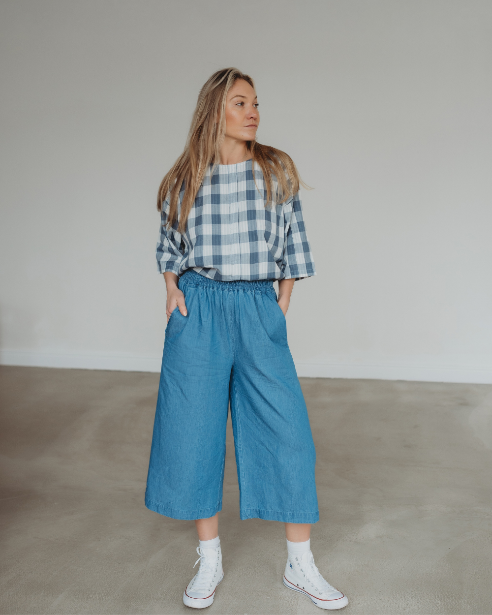 In a minimalist room, a woman wears a blue and white plaid shirt with three-quarter sleeves paired with Judy Washed Denim Blue Trousers featuring an elastic waistband. Her long blonde hair complements her white high-top sneakers as she casually rests her hands in her pockets, looking to the side.