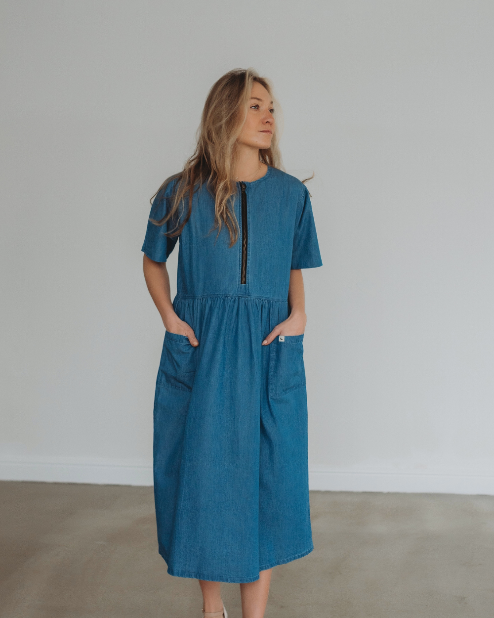 A person with long, wavy blonde hair stands against a light gray wall, wearing the Magda Washed Denim Blue Dress made from organic cotton. The dress features short sleeves and a front zipper for easy breastfeeding access, and their hands rest in its pockets.