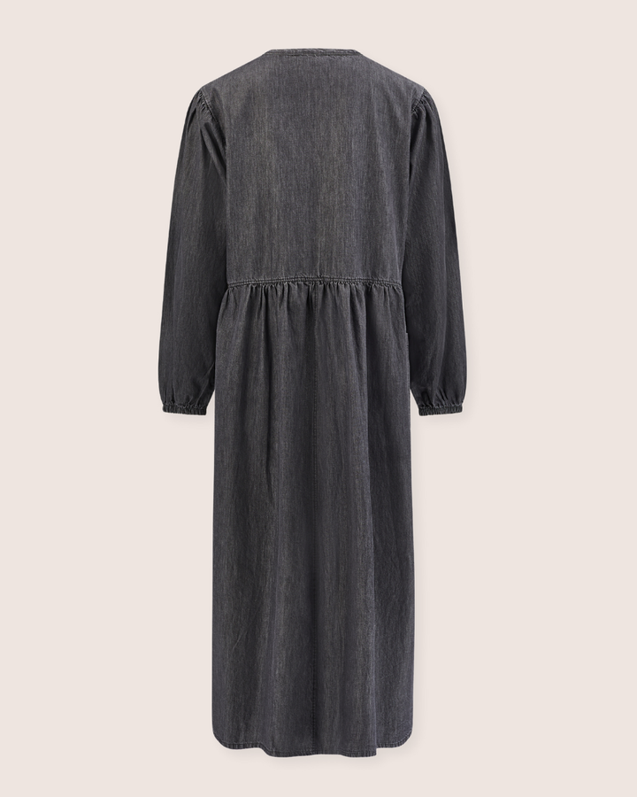 Back view of the long-sleeved Magda Black Denim Dress in dark grey, featuring a loosely gathered waist. Made from organic cotton denim, it boasts a simple design with soft texture and puffy sleeves. Background is light beige.