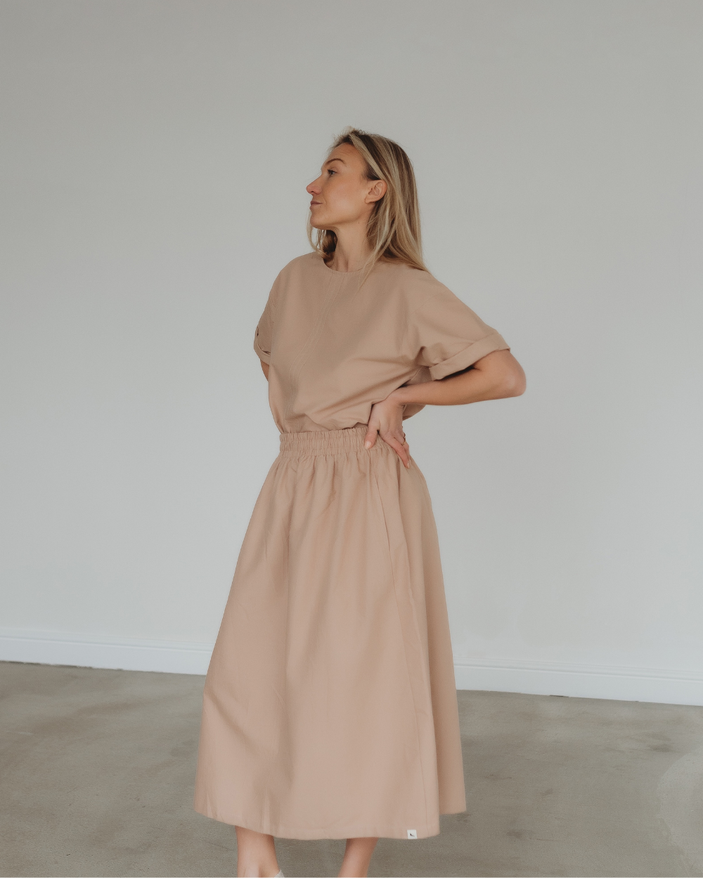 In a neutral room, a woman dons the Joyce Easy Fit Clay Skirt with an elastic waistband, paired with a beige, short-sleeved top. Her hands rest on her hips as she gazes left. This ensemble highlights a relaxed, flowy style ideal for any sustainable wardrobe.