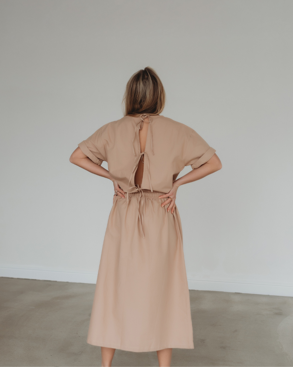 A person with blonde hair faces away from the camera, wearing a Joyce Easy Fit Clay Skirt featuring an elasticated waistband. The rooms plain white wall and gray floor enhance the skirts comfort and style.