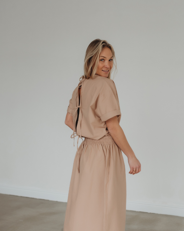 A woman with long blonde hair smiles, glancing over her shoulder. She is wearing the Joyce Easy Fit Clay Skirt, a light brown skirt tied in a bow at the open back—ideal for a sustainable wardrobe. The background features a plain, light-colored wall and floor.