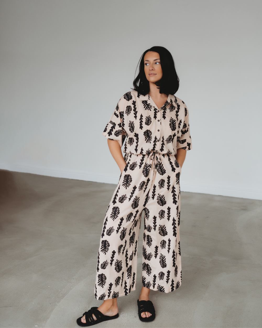 A woman with long black hair stands against a neutral background, wearing the contemporary Iris Seaweed Print Jumpsuit made of EcoVero viscose. It has black leaf patterns, a drawstring waist and pairs perfectly with her black sandals as she maintains a relaxed posture, gazing to the side.