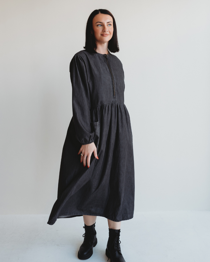 A person with shoulder-length black hair stands against a white background, wearing the Magda Black Denim Dress and black boots. They have a slight smile, with their hands relaxed by their sides.