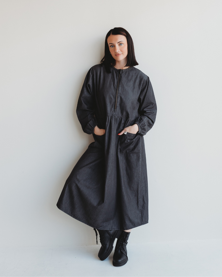 A person with shoulder-length dark hair stands against a white wall, wearing the Magda Black Denim Dress. The oversized midi dress features long sleeves, a front zipper for easy breastfeeding access, and pockets. They have a relaxed expression and are wearing black boots.