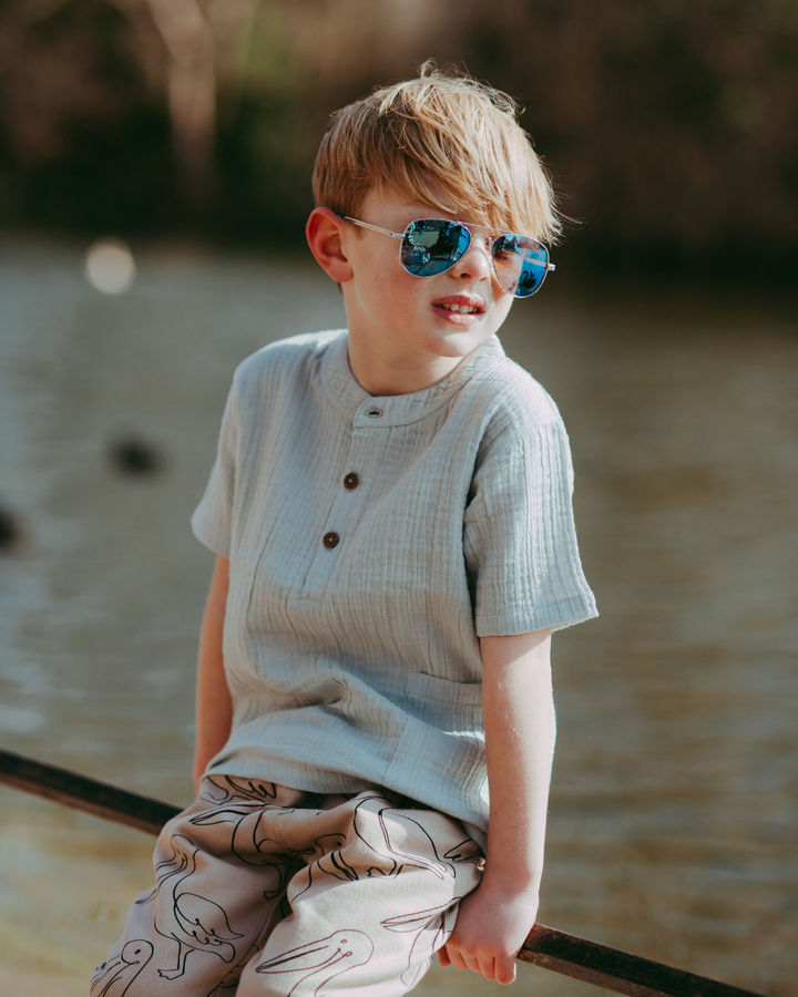 A young boy in a Sky Gauze Shirt with a mandarin collar and patterned pants sits on a railing by the lake. Wearing sunglasses and with slightly tousled hair, he enjoys the warm sunlight contributing to the relaxed outdoor scene.