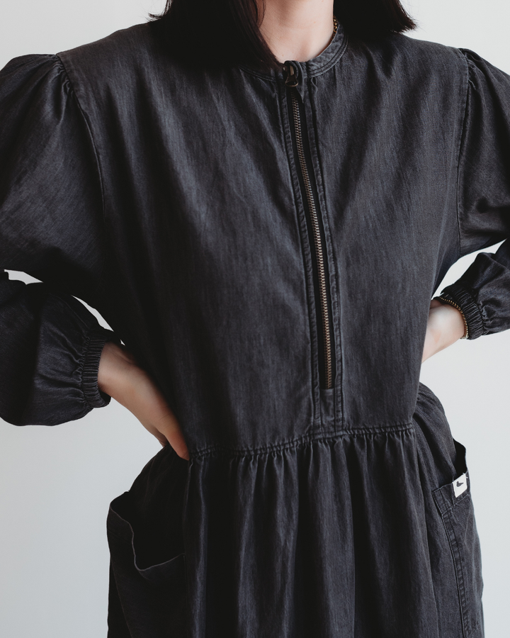 A person wearing a Magda Black Denim Dress, featuring puffed sleeves and a front zipper for easy breastfeeding, stands with hands on hips. The dress is made from organic cotton denim. The photo focuses on the upper body against a plain background.