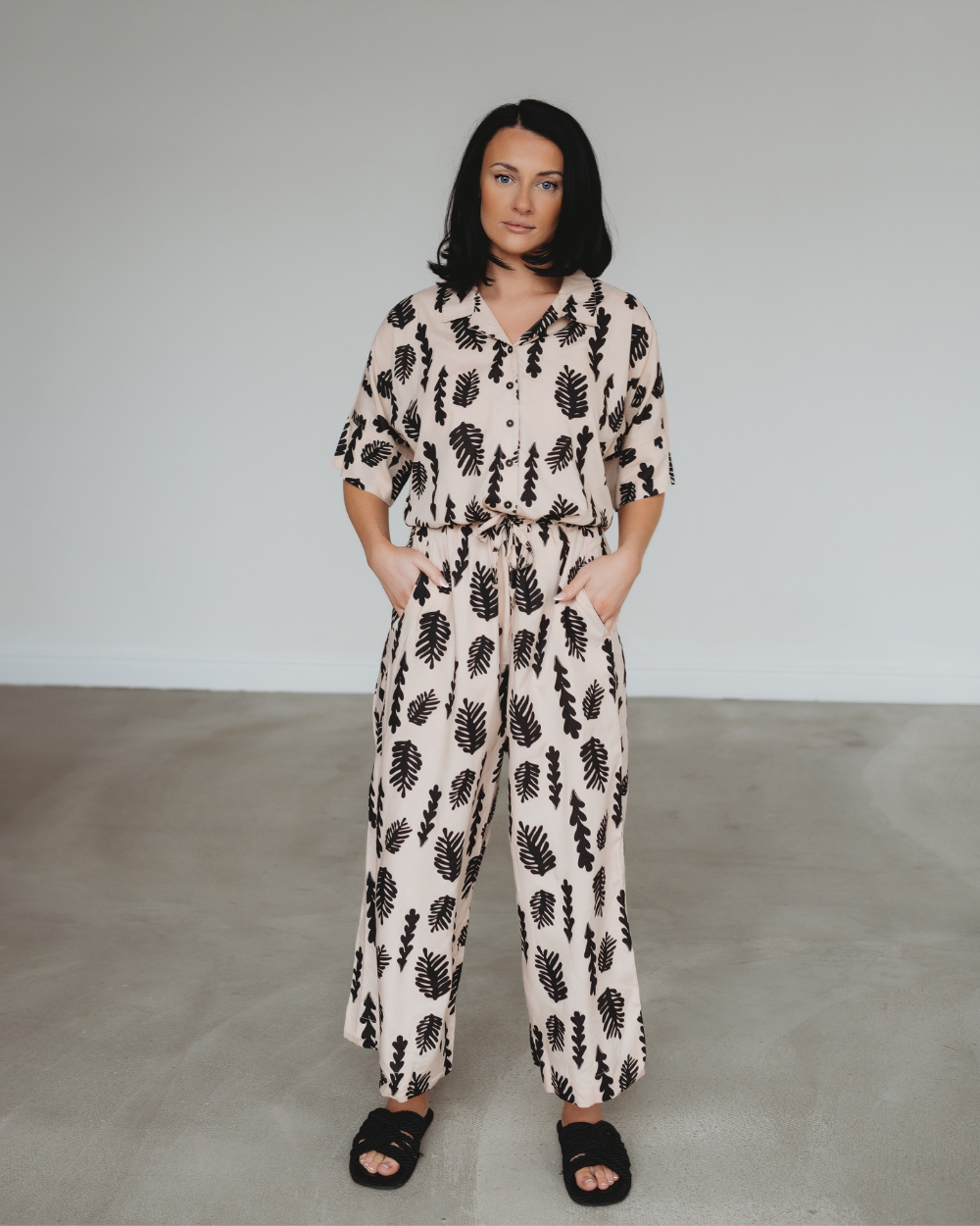 A woman stands confidently in a room, wearing the Iris Seaweed Print Jumpsuit made from EcoVero viscose with black leaf patterns. With her hands in pockets and black sandals, she exudes effortless style against a plain gray wall and floor.