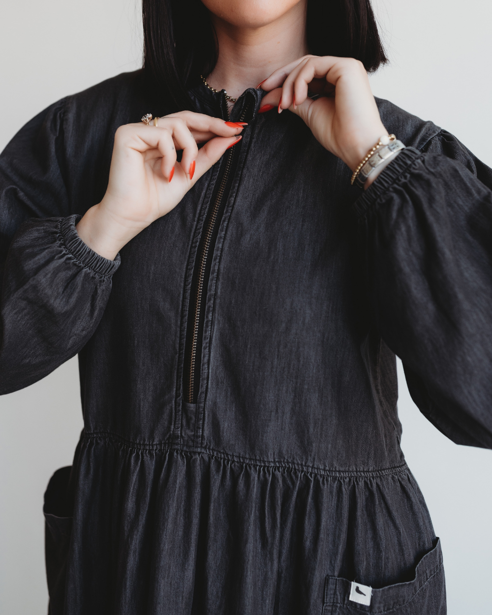 A person with dark hair zips up the Magda Black Denim Dress in organic cotton, featuring a gathered waist and puffed sleeves. Their red nails complement the bracelets and ring, while the patch pocket adds functionality.