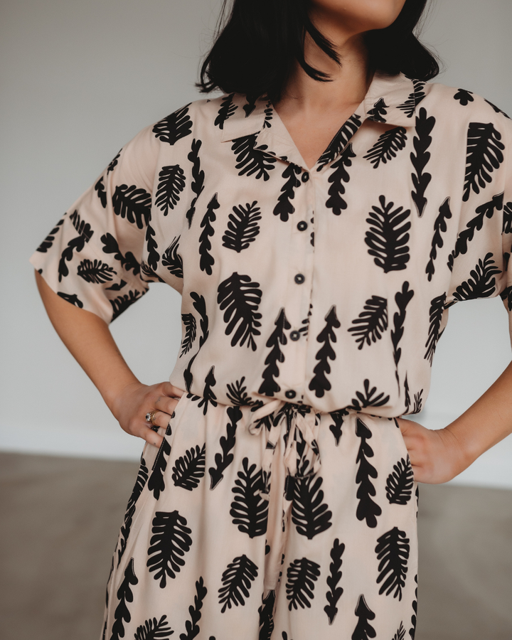 Person wearing a stylish Iris Seaweed Print Jumpsuit with black leaf patterns, posing with hands on hips. The jumpsuit is crafted from EcoVero viscose, features short sleeves, a button-down front, and a tie waist. Cropped view focuses on the outfit against a neutral background.