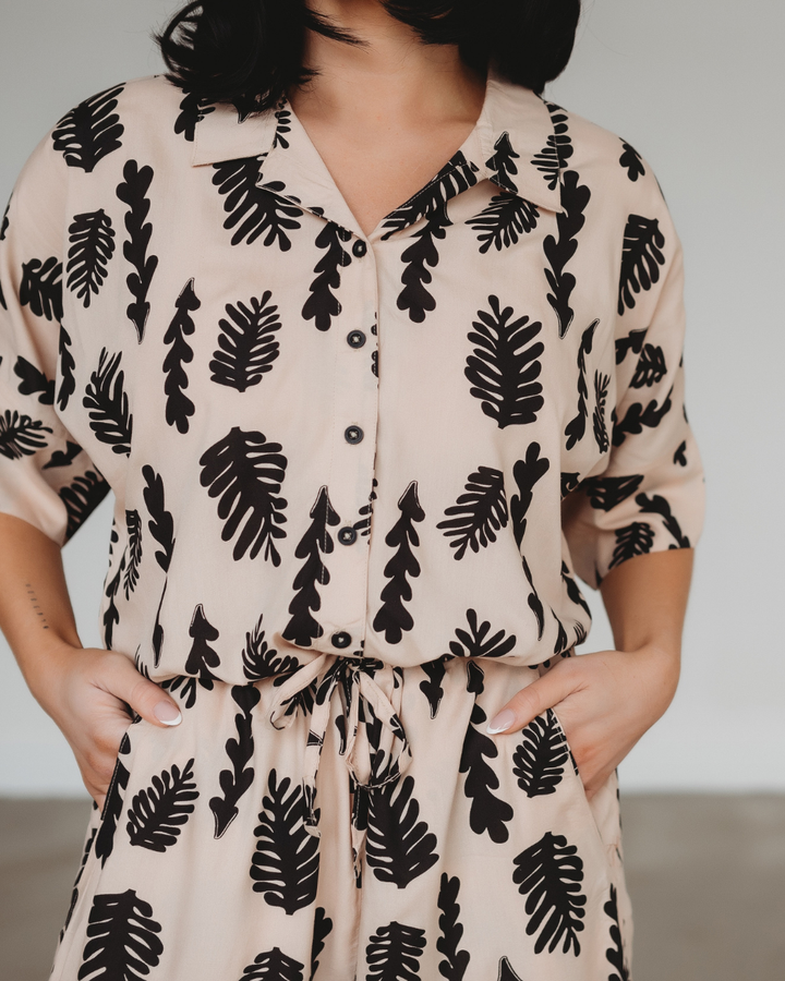 A person models the Iris Seaweed Print Jumpsuit, featuring a contemporary beige design with black leaf patterns made from EcoVero viscose. The button-up shirt and matching pants exude sophistication as they stand against a plain white background, hands casually in pockets.