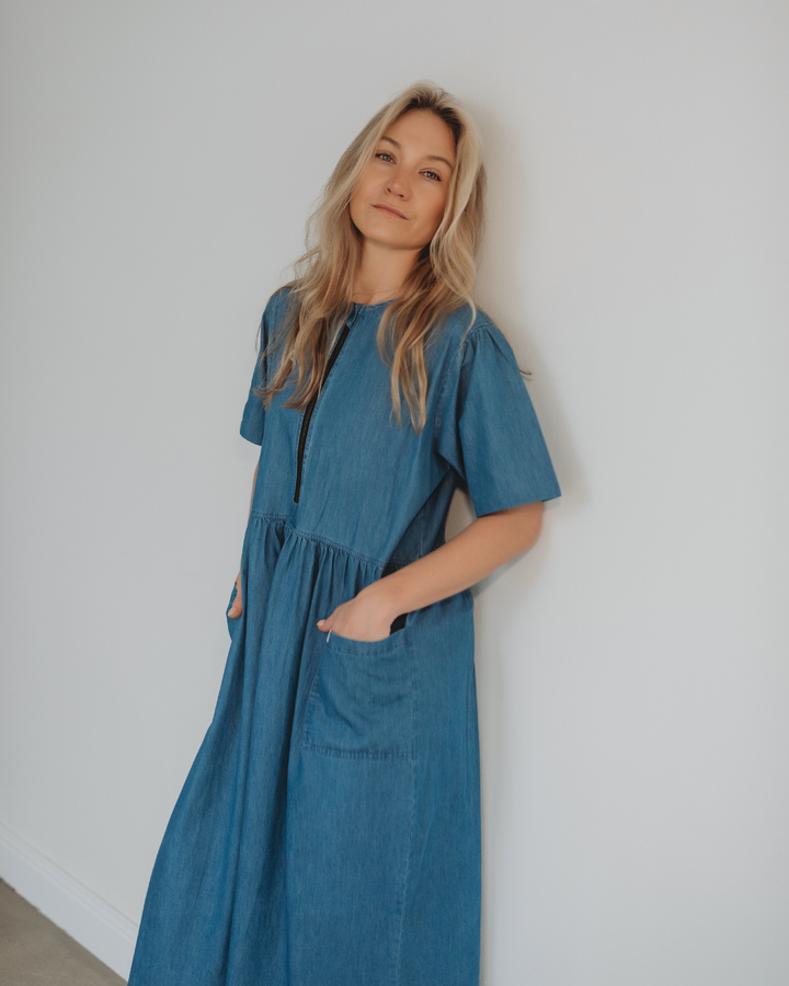 A person with long blonde hair leans against a white wall wearing the Magda Washed Denim Blue Dress made from organic cotton. With hands in pockets, they display a relaxed expression, embodying comfort and the style of sustainable fashion.