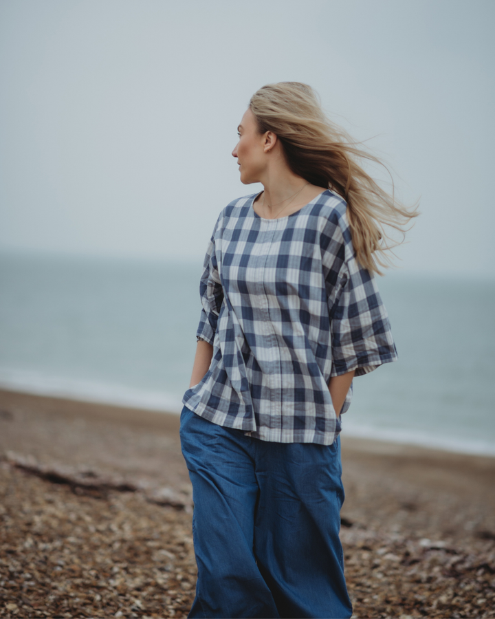 A person with long hair, in a Doris Tie Back Blue Check Top and blue pants, stands on a rocky beach with hands in pockets, gazing away at the sea under an overcast sky.