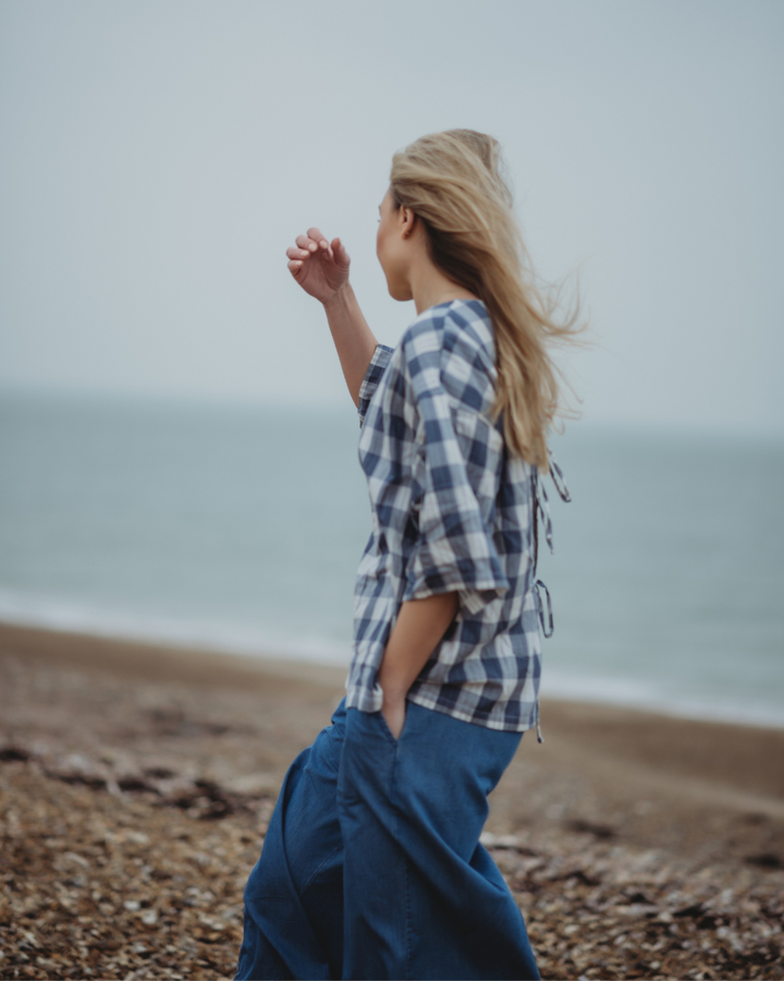 A person with long blonde hair strolls along a pebbly beach in the Doris Tie Back Blue Check Top and wide-legged organic cotton blue pants. The sea stretches in the background under a cloudy sky, creating a serene scene.