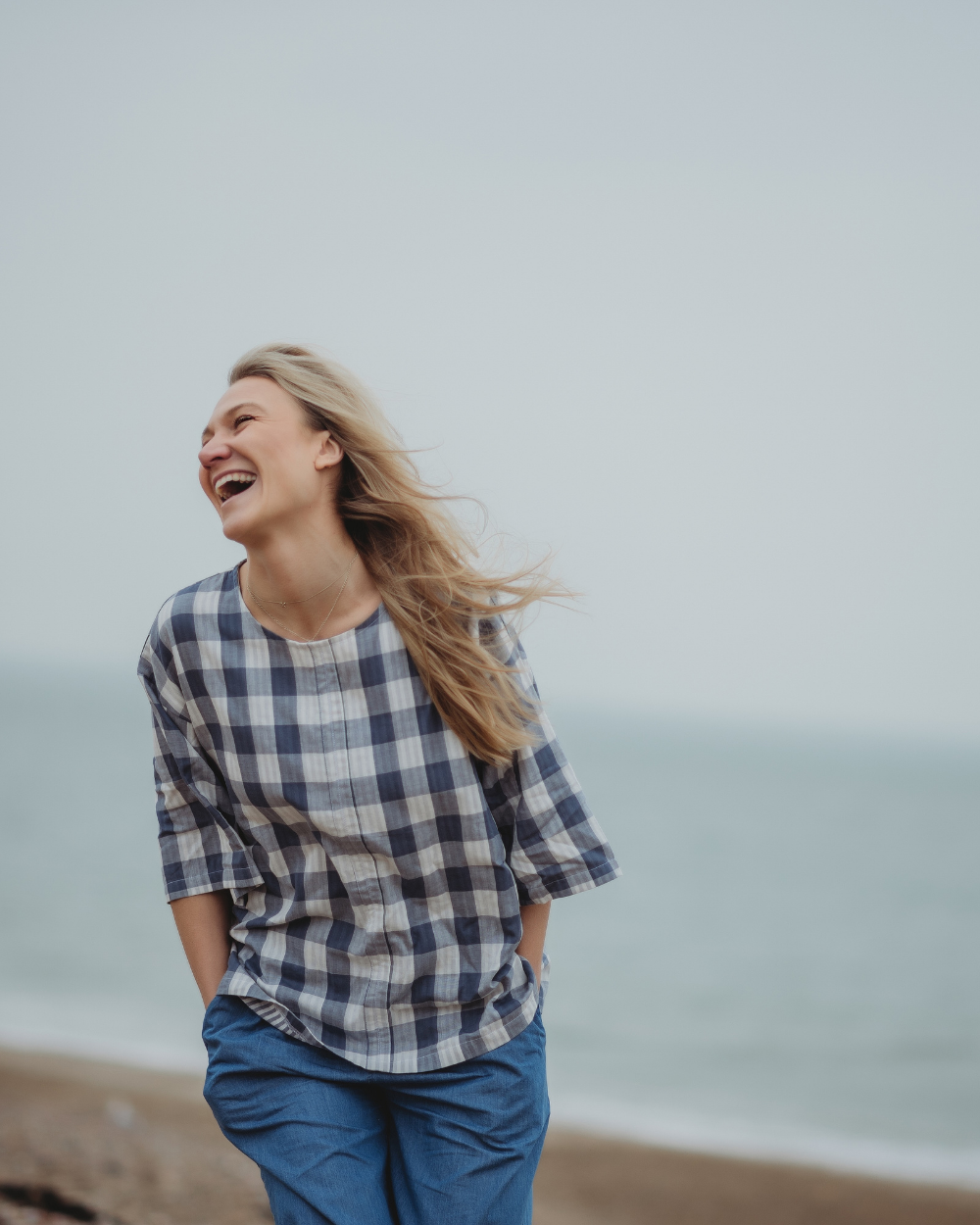 A person with long, flowing hair is joyfully laughing on a beach, wearing the Doris Tie Back Blue Check Top and blue organic cotton pants. The ocean and sky create a calm, blurred background.