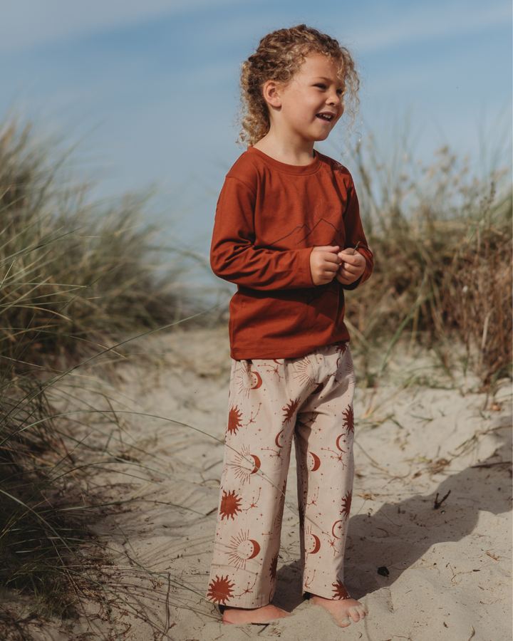 Amid tall grass on a sandy path, a curly-haired child wears the eco-friendly Wild Mountains Top in rust, crafted from organic cotton. Patterned sun and moon pants complement the look against a clear blue sky backdrop.