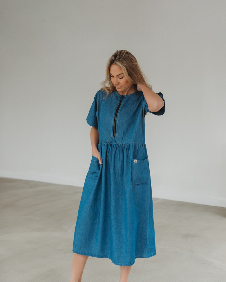 Indoors, a person gently touches their hair while wearing the Magda Washed Denim Blue Dress, made from organic cotton. The dress showcases short sleeves and handy pockets, set against a plain, light-colored wall and floor.