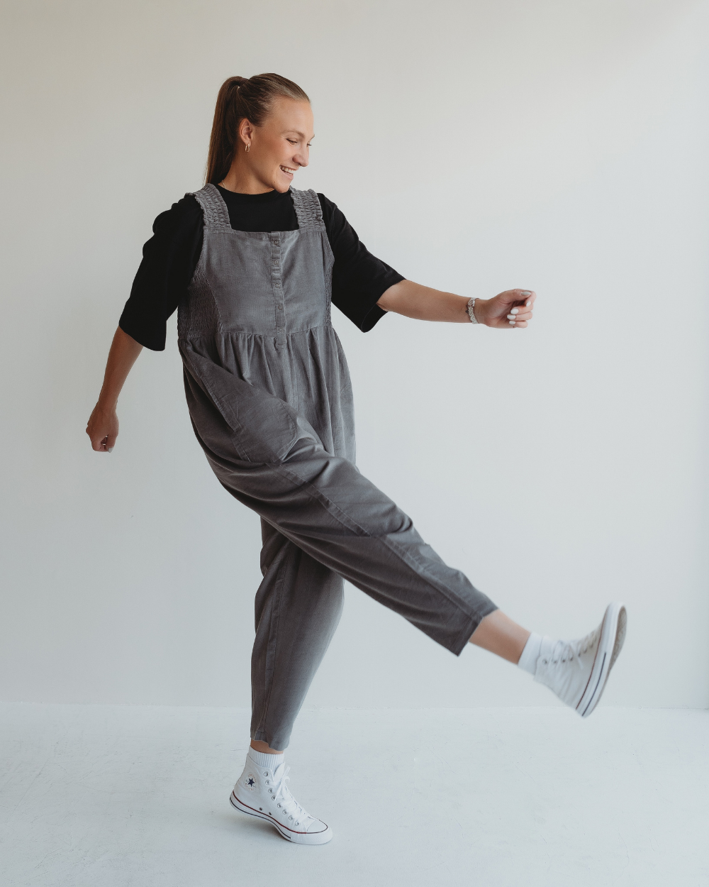 A person wearing a black top and the Alice Charcoal Jumpsuit playfully kicks a leg forward. Their long hair is tied back in a ponytail, and theyre sporting white high-top sneakers. A plain white wall background beautifully highlights the shaped silhouette.
.
