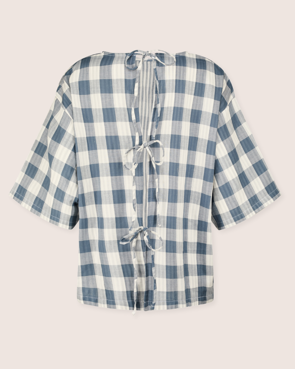 The Doris Tie Back Blue Check Top is a short-sleeved woven shirt featuring a blue and white checkered pattern. Made from organic cotton, it showcases a unique design with an open slit back tied by three bows, giving it a light, airy look ideal for casual wear.