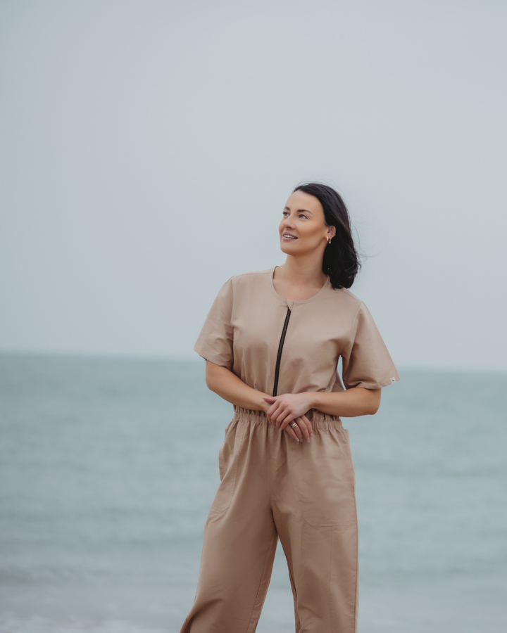 On a beach, a woman in an Ethel Plain Clay Jumpsuit gazes sideways. The ocean extends behind her beneath a cloudy sky, and the jumpsuits zip-up-and-go style offers both comfort and convenience.