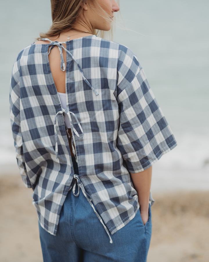 At the beach, someone wears a Doris Tie Back Blue Check Top, featuring an open back with tied strings. They have their hands in the pockets of blue pants, and their hair is loose as sand and sea fill the background.