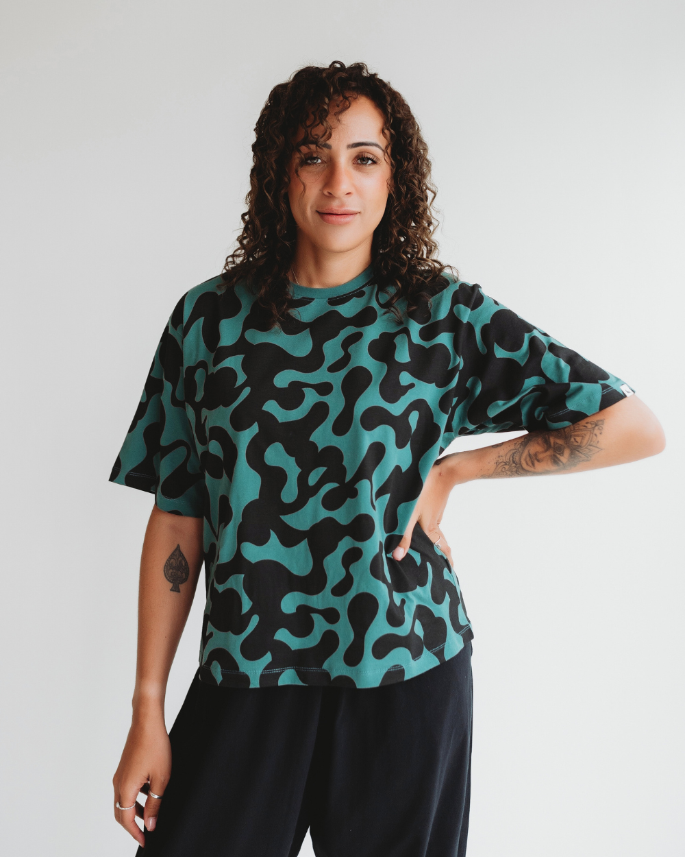 A person with curly hair wearing an Elsie Misty Print T-Shirt made of organic cotton stands against a white background, one hand on their hip and smiling slightly.