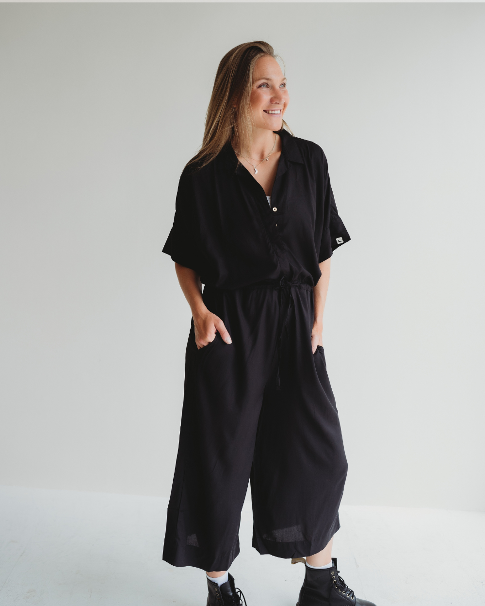 A person with long hair smiles against a plain background, wearing the Iris Black Jumpsuit with a drawstring waist and black boots. Their hands rest casually in the pockets of this stylish, sustainable EcoVero viscose outfit.