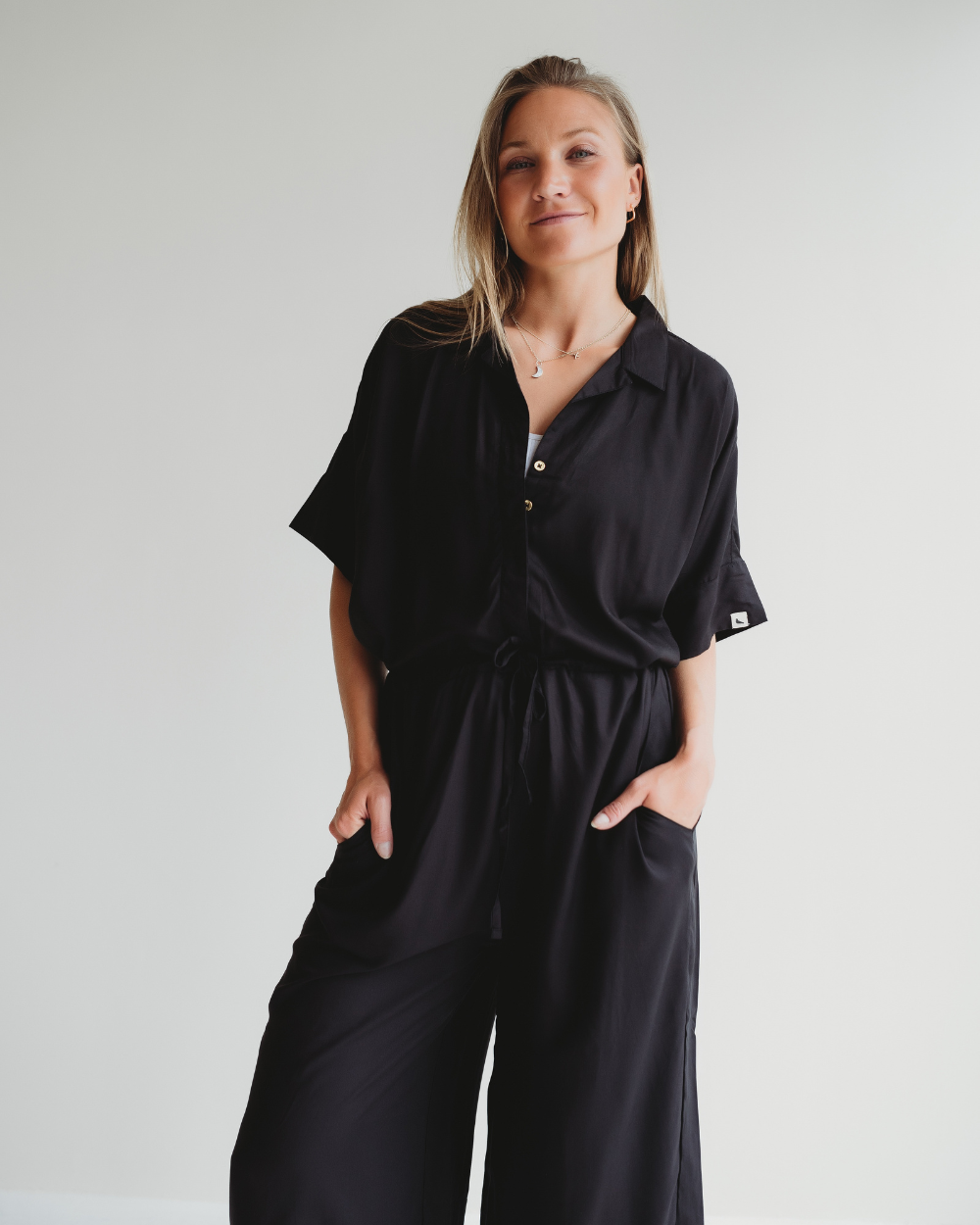 Against a plain white backdrop, someone models the Iris Black Jumpsuit, a loose-fitting attire with short sleeves and a collar. Crafted from EcoVero viscose, it boasts a drawstring waist. They casually place their hands in pockets while smiling gently at the camera.