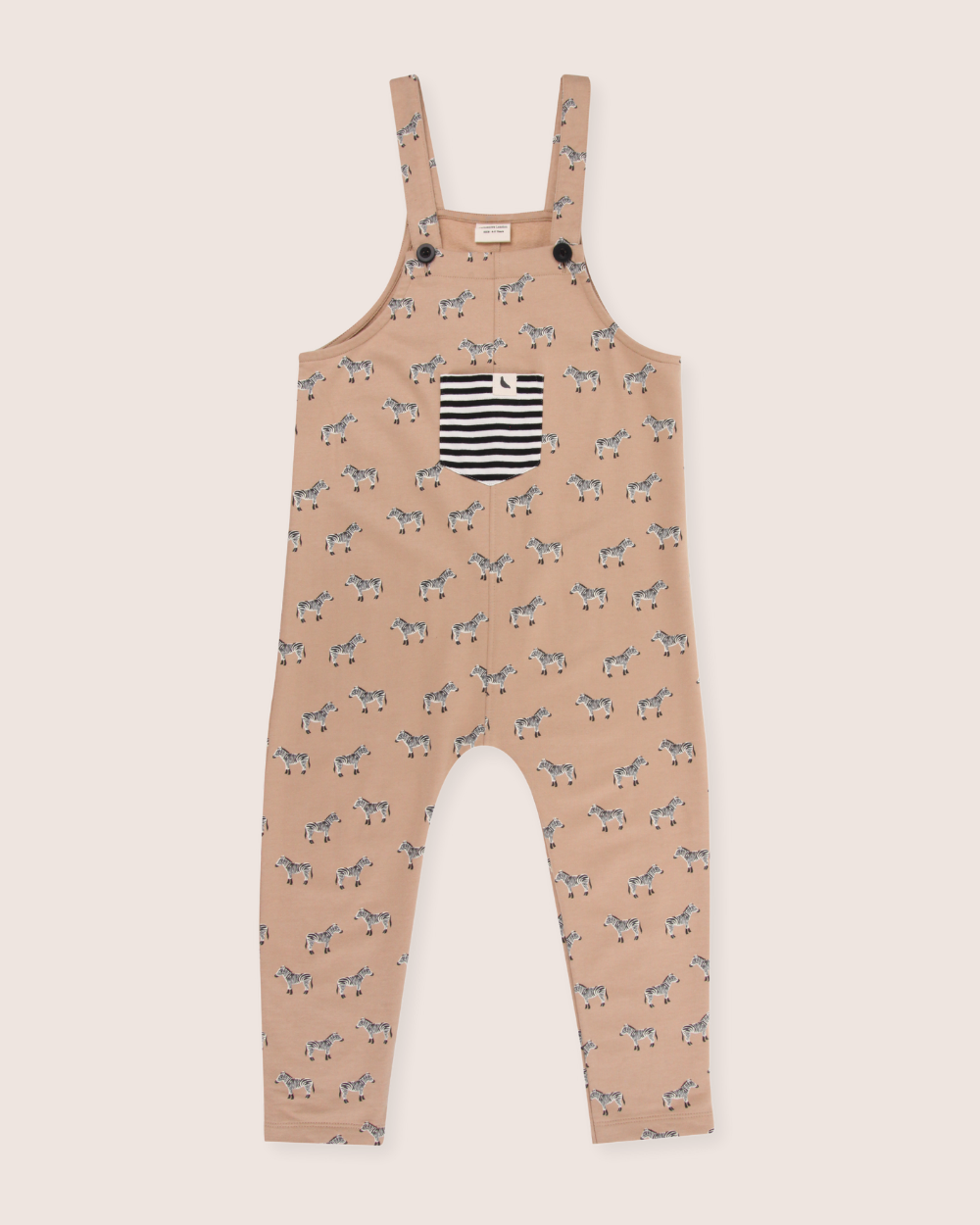 The Zebra Dungarees are eco-friendly kids overalls made from organic cotton, featuring a light brown base with a playful black and white zebra pattern. They include a front pocket with black and white stripes and button straps for casual style.